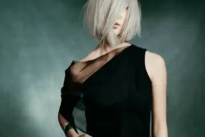 A person with chin-length, platinum blonde hair wears a one-shoulder black dress and stands against a dark background.