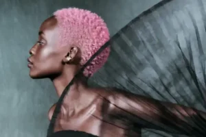 A person with short pink hair poses in profile against a gray background, draped in sheer black fabric.
